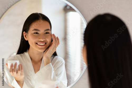 Mirror reflection of happy asian lady applying eye care product