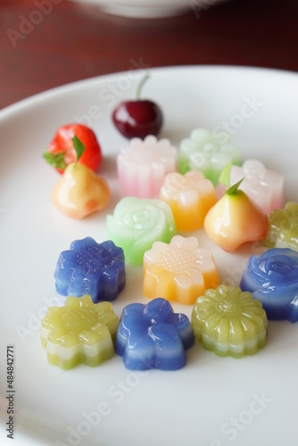 Mixed beautiful Thai dessert flower shape is Layer Sweet Cake & A fruit shape is Deletable Imitation Fruit in white plate on wooden table. (Thai name is Kanom Chan, Look Choup) #484842771