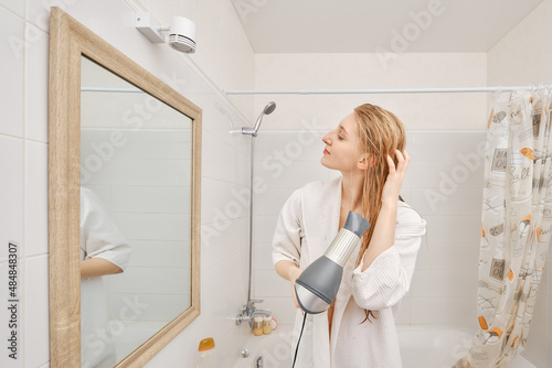 Woman in bathrobe drying her hair with dryer standing in profile in front of the mirror