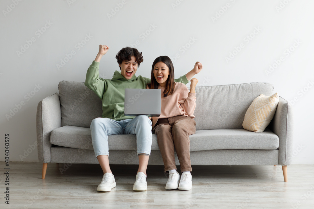 Young Asian couple with laptop gesturing YES, celebrating online business success, sitting on couch at home, copy space