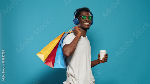 Casual man carrying shopping bags and wearing headphones. Modern person listening to music while holding handbags with clothing from retail shop and cup of coffee. Shopaholic adult