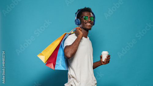 Casual man carrying shopping bags and wearing headphones. Modern person listening to music while holding handbags with clothing from retail shop and cup of coffee. Shopaholic adult photo