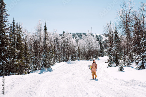 Russia. Sheregesh. Girl snowboarder in black sun goggles in winter in sunny weather outdoors among the Christmas trees and snow. The girl goes with a snowboard in her hands, smiling and posing. 