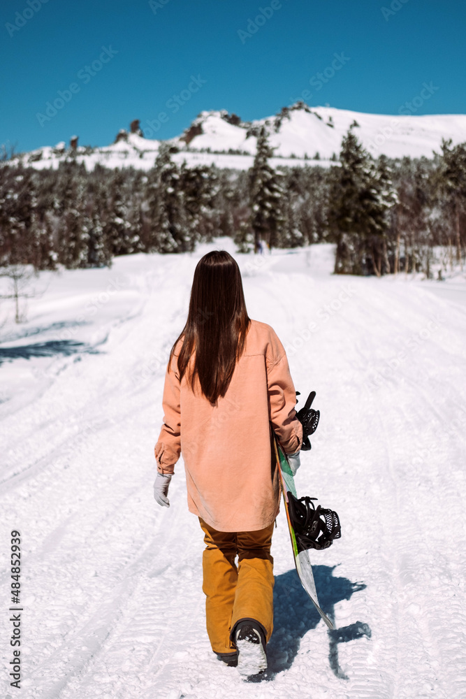 Russia. Sheregesh. Girl snowboarder in winter in sunny weather outdoors among the Christmas trees, mountains and snow. The girl goes with a snowboard in her hands, turned back to the camera.
