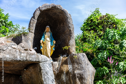 Statue of Virgin Mary in a rock cave chapel outside the St Anne’s Catholic Church in Baie Ste Anne, Praslin, Seychelles, with tropical vegetation around. photo