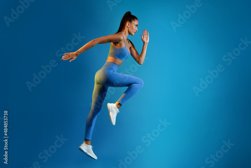 Fototapeta Naklejka Na Ścianę i Meble -  Sporty Woman Exercising Doing Elbow-To-Knee Crunch Standing On Blue Background