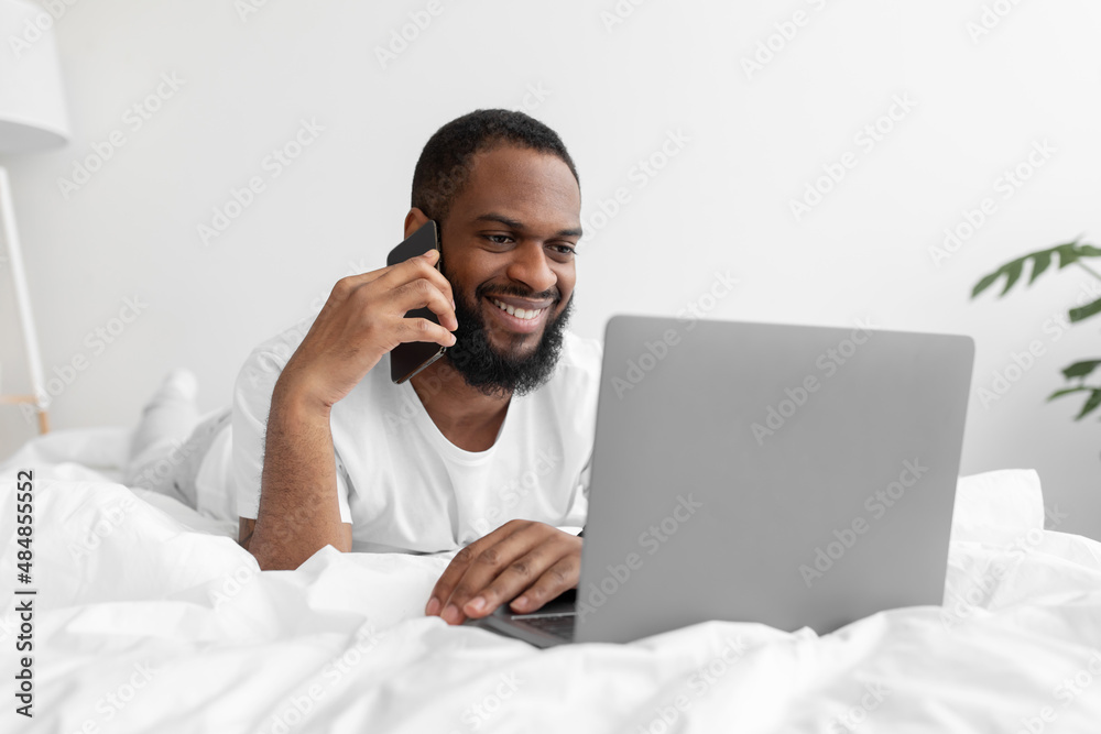 Smiling young black man with beard speaks by phone, looks at laptop on white bed in bedroom interior Smiling young black man with beard speaks by phone, looks at laptop on white bed in bedroom interior