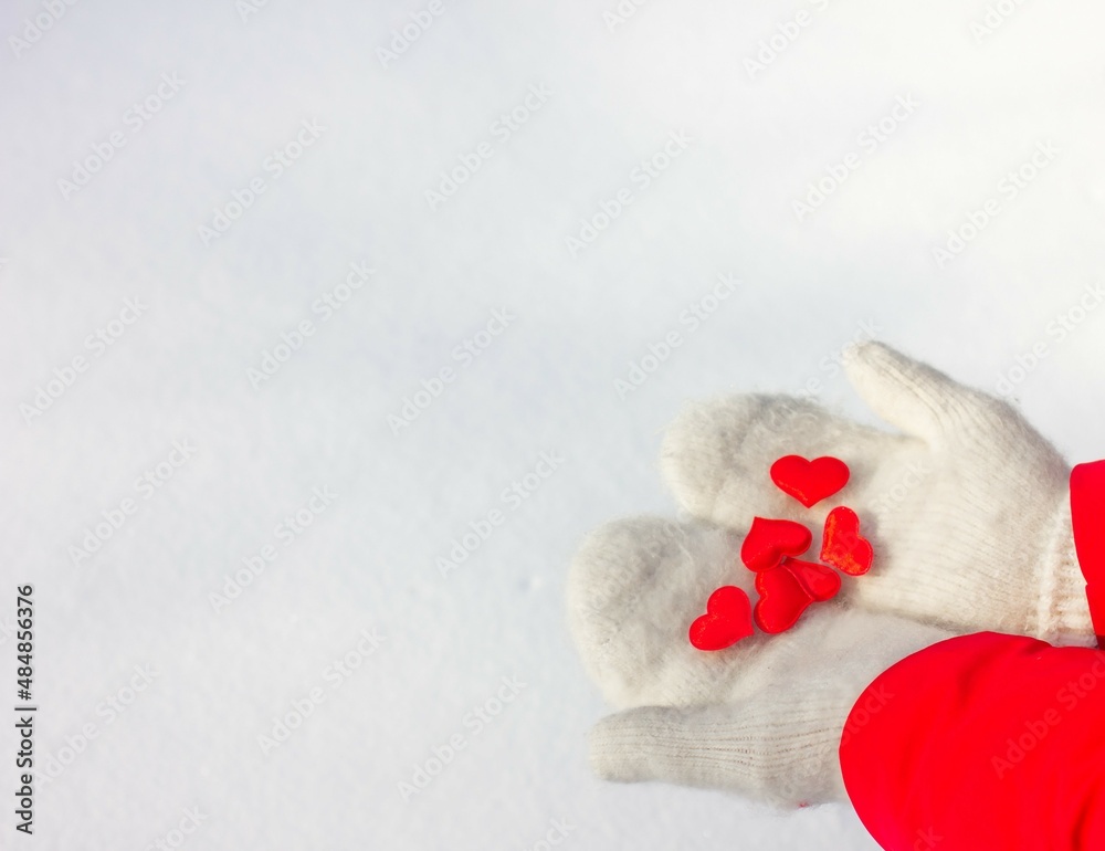 Fototapeta premium Female hands in knitted white mittens hold a silk red heart on a snowy background. Concept of love and valentine.