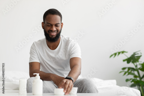 Cheerful attractive young african american man with beard takes jar of cream in hand in bedroom interior