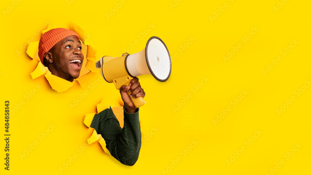 Emotional young black guy shouting into megaphone through hole in ripped orange paper, banner with free space Emotional young black guy shouting into megaphone through hole in ripped orange paper, banner with free space