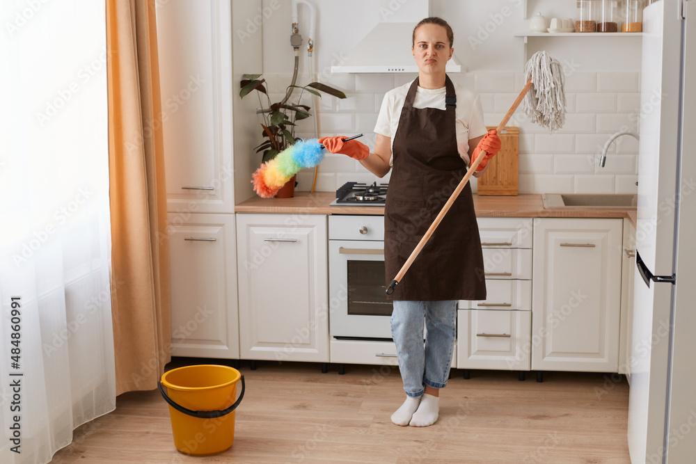 Indoor shot of bored young woman housekeeper holding brush and ppduster, washing her kitchen, wearing brown apron, jeans and white t shirt, standing and looking at camera. Indoor shot of bored young woman housekeeper holding brush and ppduster, washing her kitchen, wearing brown apron, jeans and white t shirt, standing and looking at camera.