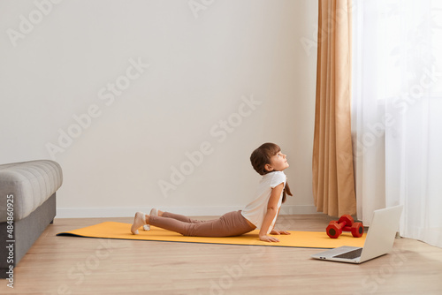 Indoor shot of little cute girl practicing yoga pose on a mat indoor, stretching her body, wearing casual clothing, having braids, posing in light living room near window.