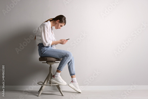 Woman with bad posture using smartphone while sitting on stool near light grey wall indoors, space for text