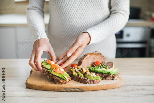 Detail of young woman hands making healthy meal of avocado toast and vegetables, indoor natural light, bokeh background