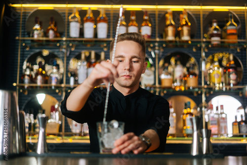 Portrait of bartender male stirring ice cubes in glass using spoon standing on bar counter, on background of shelves with different alcoholic drinks. Barman making cocktail in bar with dark interior.