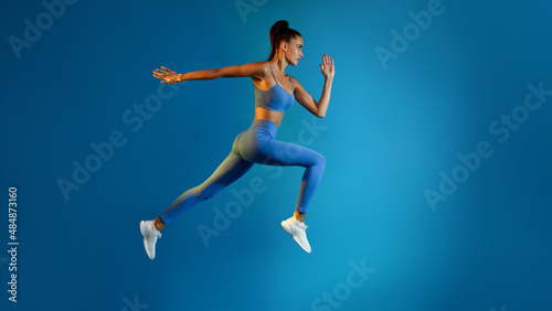 Determined Young Sportswoman Jumping Posing In Mid-Air On Blue Background © Prostock-studio