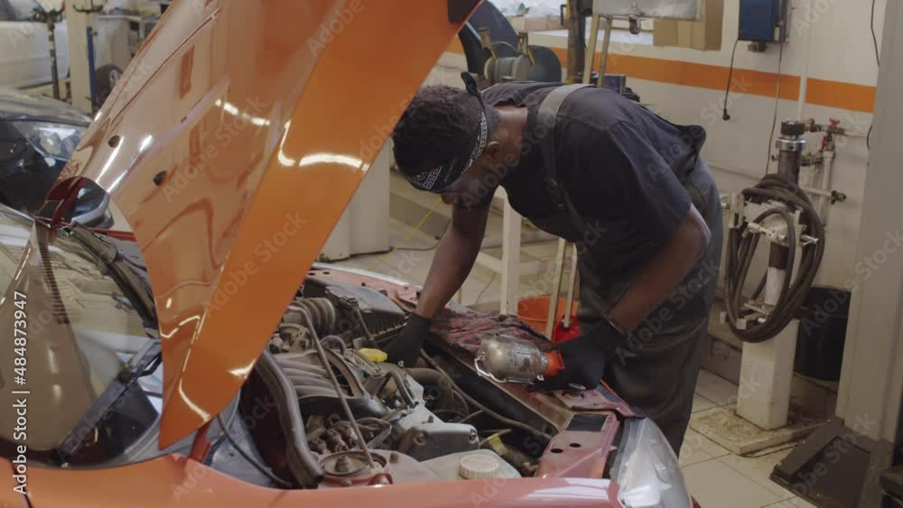 African American mechanic in uniform and gloves examining car parts under hood with lamp while working in auto repair shop