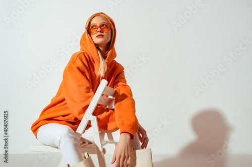 Fashionable girl wearing orange hoodie, glasses looking up,  posing on white background. Copy, empty space for text