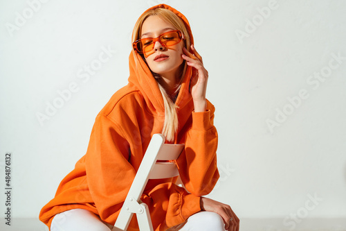 Indoor fashion portrait of young confident blonde woman wearing trendy orange hoodie, color sunglasses, posing on white background. Copy, empty space for text photo