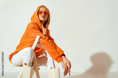 Young confident blonde girl wearing trendy orange hoodie, color sunglasses, posing on white background. Studio fashion portrait. Copy, empty space for text photo