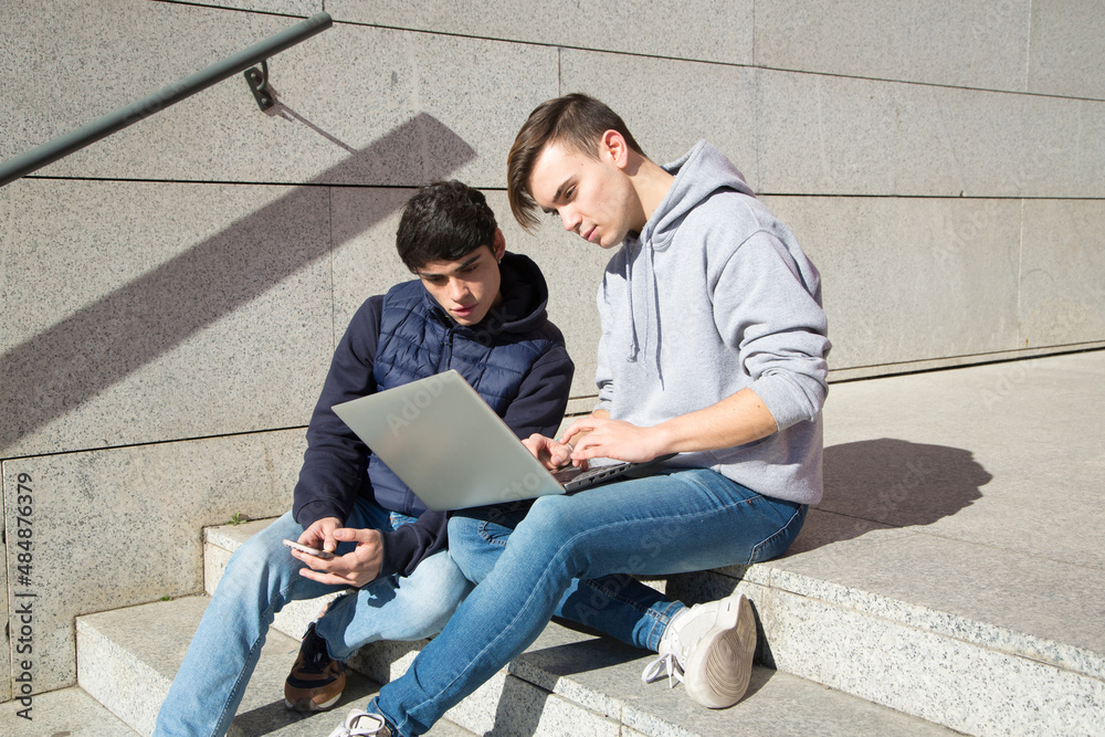 two young male university students sitting on a staircase looking at a laptop computer
