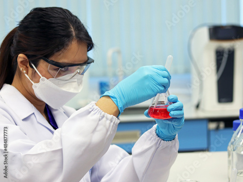 The scientist wears PPE and holds an Erlenmeyer flask laboratory, with red solvent solution from reaction methyl orange indicator acid titration experiment, analysis compounding in wastewater sample.