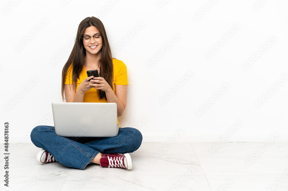 Young woman with a laptop sitting on the floor sending a message with the mobile Young woman with a laptop sitting on the floor sending a message with the mobile