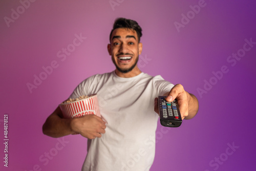 Cheerful millennial Arab guy watching TV and eating popcorn, switching channels with remote control in neon light