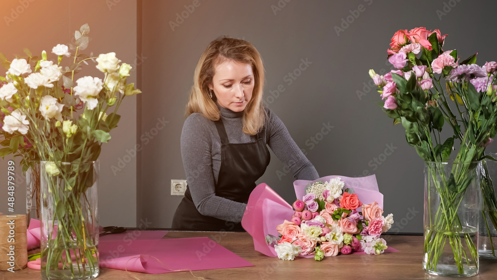 Florist woman in black apron makes elegant wedding bouquet with colorful roses wrapping in pink paper on wooden table in floristry studio, sunlight Florist woman in black apron makes elegant wedding bouquet with colorful roses wrapping in pink paper on wooden table in floristry studio, sunlight