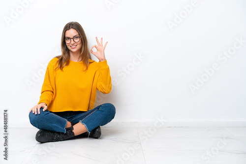 Young caucasian woman sitting on the floor isolated on white background showing ok sign with fingers