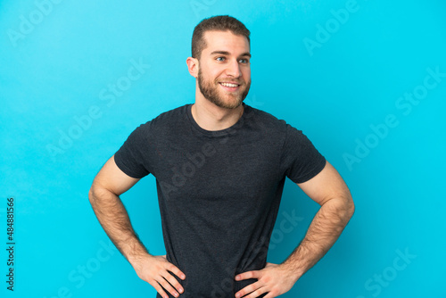 Young handsome caucasian man isolated on blue background posing with arms at hip and smiling