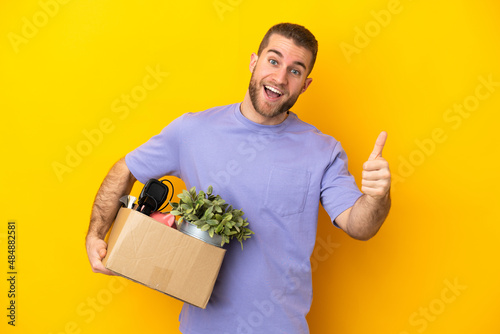 Young caucasian making a move while picking up a box full of things isolated on yellow background with thumbs up because something good has happened