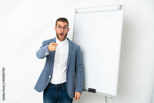 Young handsome caucasian man isolated on white background giving a presentation on white board and surprised while pointing front