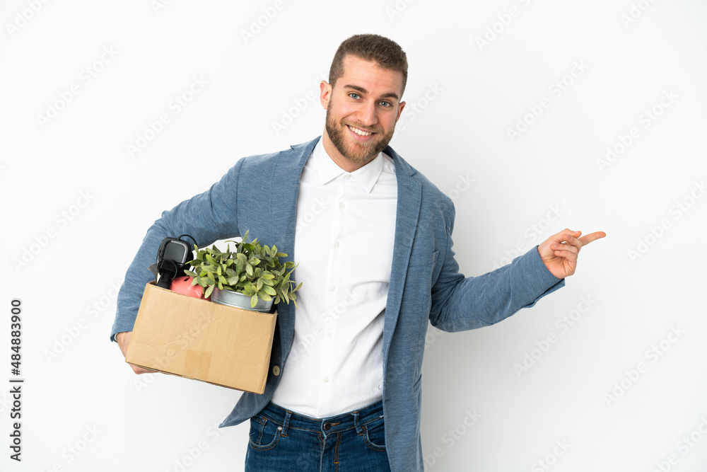 Young caucasian making a move while picking up a box full of things isolated on white background pointing finger to the side Young caucasian making a move while picking up a box full of things isolated on white background pointing finger to the side