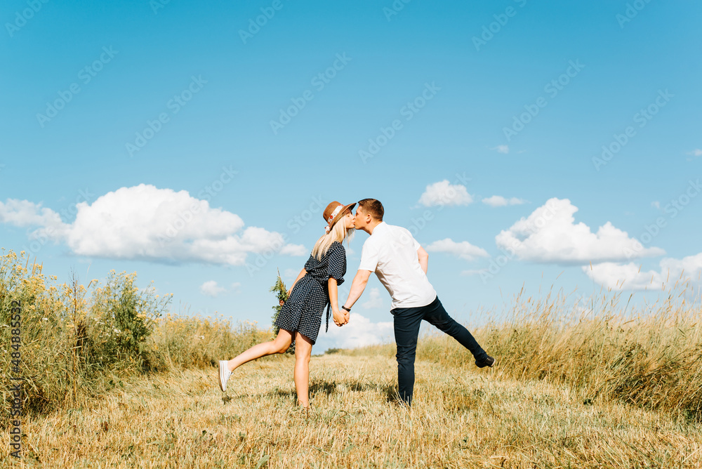 Romantic date, valentine's day, love story concept. Young caucasian couple in love kissing in field on sunny summer day, outdoors. Back view of flirting man and woman holding hands together in meadow Romantic date, valentine's day, love story concept. Young caucasian couple in love kissing in field on sunny summer day, outdoors. Back view of flirting man and woman holding hands together in meadow