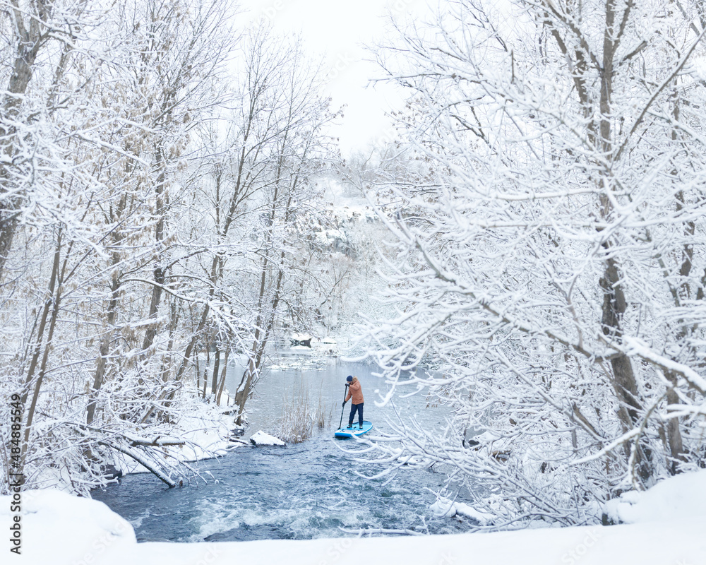 a man is engaged in SUP boarding in winter on the river in a picturesque place where everything is covered in snow
