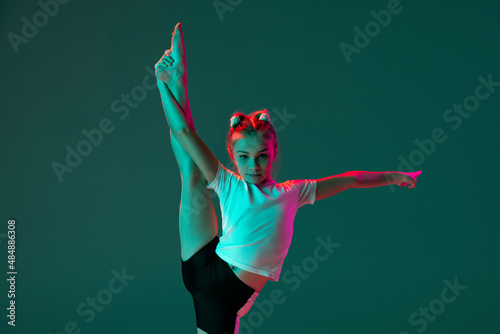 Little flexible girl, rhythmic gymnastics artist training isolated on green studio background in neon pink light. Grace in motion, action. Doing exercises in flexibility.