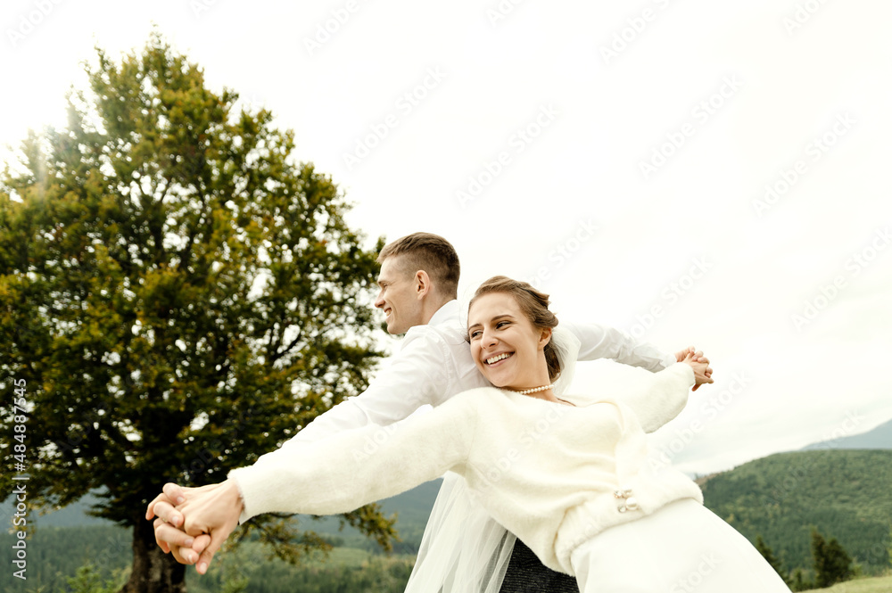 cheerful bride and groom stand with their backs to each other and hold hands on the top of the mountain. wedding day of a young couple in love cheerful bride and groom stand with their backs to each other and hold hands on the top of the mountain. wedding day of a young couple in love