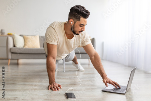 Arab guy using laptop and smartphone, listening to music during workout, communicating to personal trainer on webcam