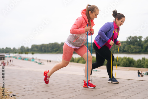 Side view of motivated fat young woman doing squats exercises using fitness tape for weight loss with personal trainer outdoor in summer day. Overweight female with coach stretching before running.