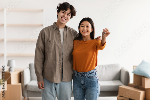 Portrait of young happy Asian couple holding house key, posing in apartment with carton boxes. Moving day concept