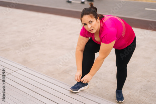 High-angle view of fat attractive young woman tying shoelaces on bench before start running, looking at camera. Sportive obese female preparing to jogging in city park for lose weight.