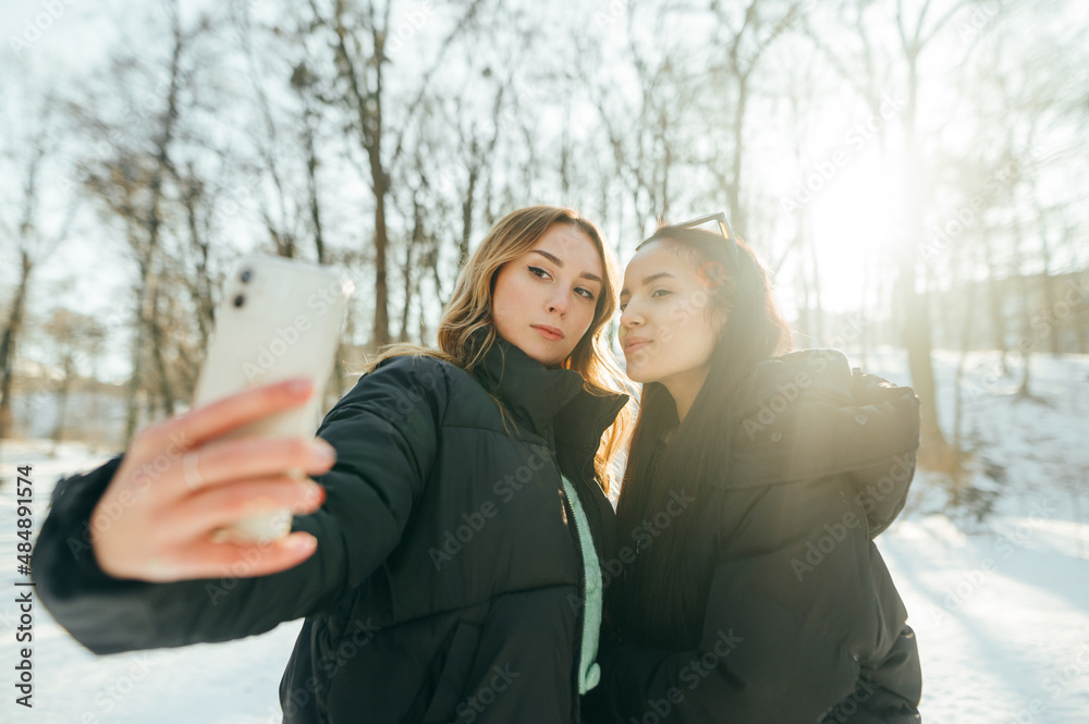 Two beautiful women take a selfie on a walk in the park on a winter snowy day, looking at the smartphone camera on a background of beautiful views. Two beautiful women take a selfie on a walk in the park on a winter snowy day, looking at the smartphone camera on a background of beautiful views.