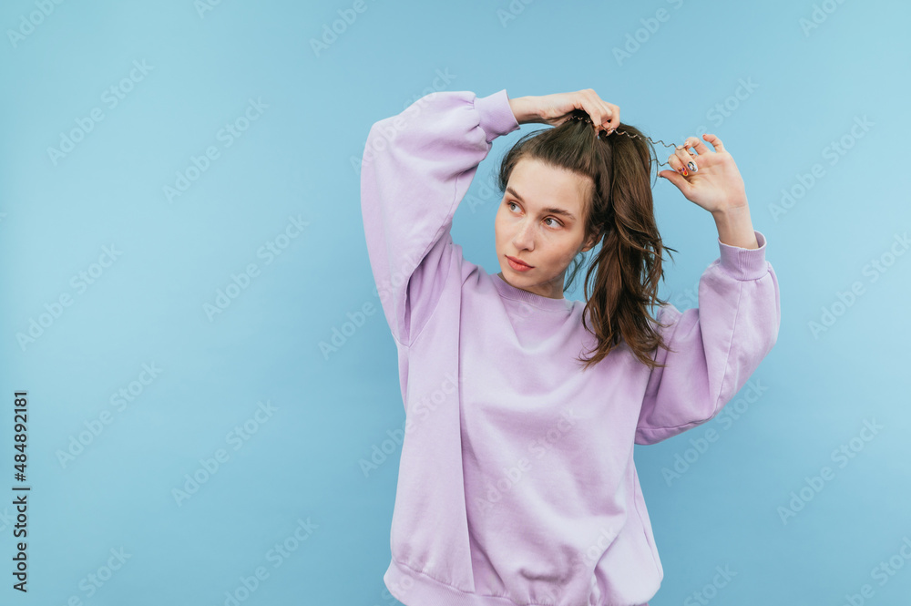 Attractive girl in a sweatshirt ties a ponytail in her hair with a serious face on a blue background and looks away at copy space Attractive girl in a sweatshirt ties a ponytail in her hair with a serious face on a blue background and looks away at copy space