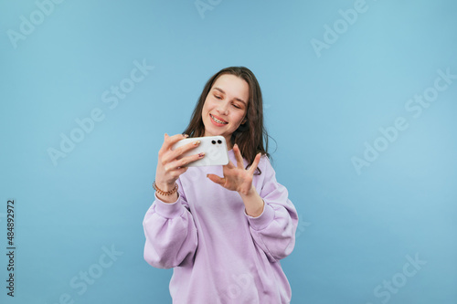 Attractive girl in a purple sweatshirt stands with a smartphone in his hands on a blue background and plays mobile games on a smartphone with a smile on his face.