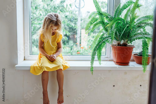 Bild auf Leinwand Sad little blonde girl in yellow dress sitting on window sill looking down near plant fern in clay pot