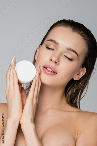 young woman with wet hair and closed eyes holding cosmetic cream isolated on grey.