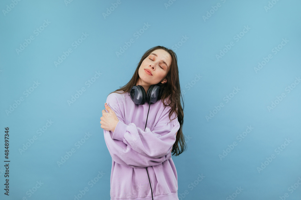 Beautiful girl in a pink sweatshirt and headphones around her neck from the cold hugs herself with closed eyes on a blue background Beautiful girl in a pink sweatshirt and headphones around her neck from the cold hugs herself with closed eyes on a blue background