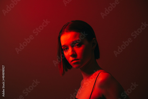 Close fashion portrait of attractive woman in red neon light on wall background, posing at camera.
