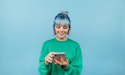 Positive lady with blue hair and green sweatshirt enjoying smartphone on blue background with smile on face looking at screen.